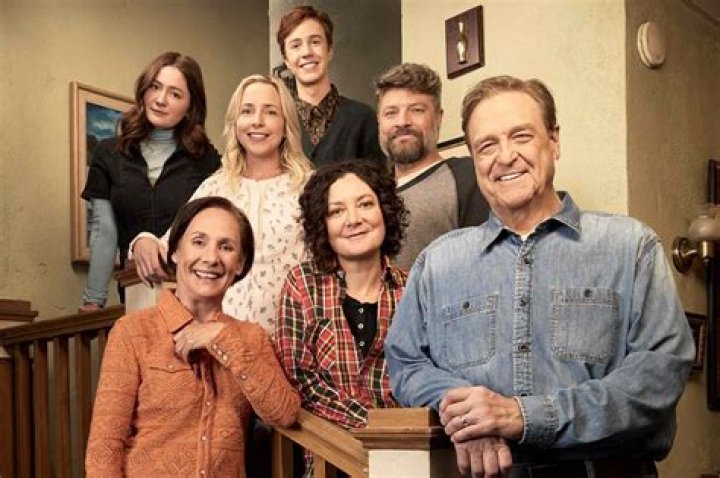The cast of The Conners sit on the couch beside an image of Katey Sagal's Louise and John Goodman's Dan embracing on their wedding day